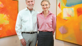 A man and a woman are standing side by side in an art gallery in Mill Valley, smiling. The gallery walls display vibrant, Expressionist paintings dominated by warm colors like orange, yellow, red, and some cooler tones like blue and white. The floor is wood, and the lighting is bright.