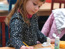 A young girl with long blonde hair wearing a black and white patterned sweater is sitting at a table engaged in a painting activity. She is dipping a paintbrush into a bowl of blue water, with art supplies scattered around her on the table—a perfect cure for cabin fever.
