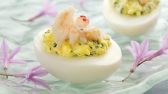 Close-up of two deviled eggs topped with shrimp and garnished with chopped herbs, placed on a clear, textured glass plate. The plate is adorned with small purple flowers, and the background is a light blue surface – a delightful recipe twist you must try!