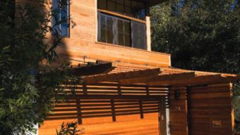 A modern two-story wooden house with large windows and a flat roof, exuding contemporary style and surrounded by trees. The front yard features a pebble and stone pathway leading to the entrance, while a wooden garage door is located on the ground level. This home embodies sustainable living under a clear, blue sky.