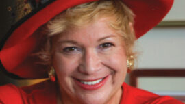 A smiling woman with blonde hair, reminiscent of Jan Wahl's signature style, is wearing a vibrant red hat adorned with decorative accents and matching red clothes. She also has gold earrings. The background features a blurred interior setting.