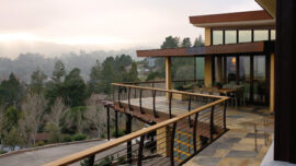 Modern house with large windows and a patio featuring an outdoor dining area. The patio overlooks a hilly landscape with a mix of trees and houses in the distance, under a misty sky. A wooden-ramped walkway extends from the patio, providing a scenic viewing point. The home boasts eco-friendly roofing, blending seamlessly with nature.