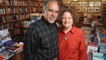 A man and a woman standing closely together, smiling at the camera, in a cozy bookstore. The background features shelves filled with various books and a table stacked with more books, including several marked as 2008 Editors' Choice. Warm lighting hangs from the ceiling, illuminating the scene.