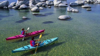 Two people kayaking in translucent, shallow, calm water among smooth rocks. They are wearing life vests, one in a red kayak and the other in a blue kayak. The background features serene Lake Tahoe and distant mountains under a clear, blue sky—an ideal setting for Easy Escapes.