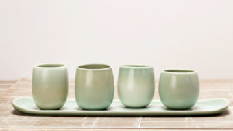 A minimalist ceramic set features four light green cups placed on an elongated matching tray. The set sits on a rustic wooden surface, and the background is a plain, light color, highlighting the simplicity and elegant design of the ceramics, flowing smoothly like gentle currents.