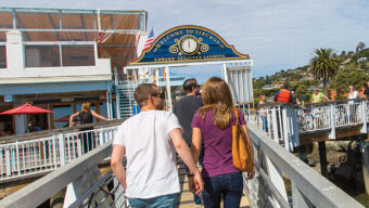Two people, one in a white shirt and blue jeans and the other in a purple shirt and dark jeans, walk hand-in-hand on a sunny day along a Marin pier towards a colorful sign that says, "Welcome to the Santa Cruz Wharf." The sky is blue with scattered clouds.