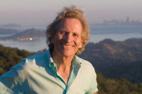 A smiling person with curly hair in a light green shirt stands outdoors against a backdrop of hills and a body of water with a distant city skyline. The clear, sunny weather adds to the scene, reminiscent of one Doug McConnell might explore.