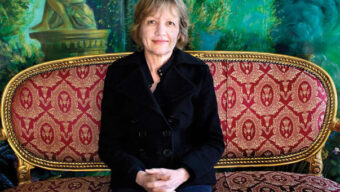 A woman with short hair and a warm smile sits on an ornate red and gold vintage sofa, ready for a lively conversation. She wears a black jacket and clasps her hands in her lap. The background features lush, green, and abstract nature-themed artwork.