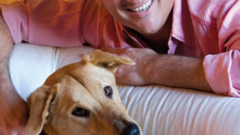 A smiling man in a pink shirt is lying on a white couch, leaning towards a light brown dog with upright ears. Mark Pitta has his hand on the dog's back, and they both appear relaxed and content.