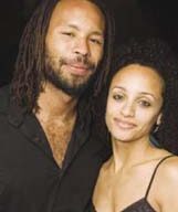 A man with shoulder-length dreadlocks and a neatly trimmed beard, dressed in a black shirt, stands next to a woman with curly hair wearing a spaghetti strap top. They are both smiling and standing closely together, each holding a sake drink.