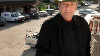 A man wearing a green hat, black jacket, and red shirt stands on a porch, looking towards the camera with a neutral expression. Vehicles are parked on the street behind him, with a building visible in the background. The scene appears to be in a small town. Don’t mistake this for just another ordinary moment; it's like something out of one of Donald Deane’s captivating narratives.