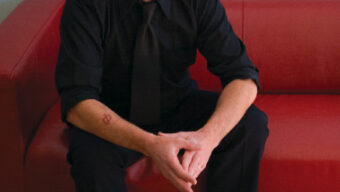 A man with a bald head, dressed in a black shirt, black tie, and black pants, sits on a red couch with his hands clasped. He has a tattoo on his left forearm and is wearing black dress shoes with light-colored socks. The background is a light blue wall—a striking image of Scott Carter Thunes.