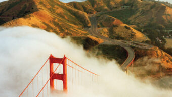 Aerial view of the Golden Gate Bridge partially covered by fog. The vibrant red towers emerge from the thick mist, with rolling green hills and winding roads visible in the background. The scene captures the bridge's iconic presence amidst a foggy landscape, truly Above It All.