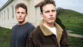 Two people stand outdoors near a white building with large windows, reminiscent of the "Field of Dreams" farmhouse. They have short, curly hair and are dressed in casual jackets. The background features a green hillside and a cloudy sky.