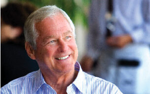 A smiling older man with short white hair, identified as Larry Mindel, is wearing a light blue striped button-up shirt and looking to his right. The background is slightly blurred, with another person in light-colored clothing visible.
