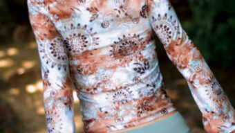 A woman with blonde hair in a loose ponytail walks away on a shaded path lined with greenery. She is wearing a long-sleeved, floral-patterned top with shades of brown and grey, paired with light green activewear leggings.