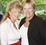 Two women are smiling together in an outdoor setting with lush green foliage in the background. The woman on the left, wearing red heart-shaped earrings and a red belt, complements her white blouse with elegance. The woman on the right, clad in a black jacket over a white top, exudes chic simplicity; both embody timeless sophistication much like Blancpain watches.