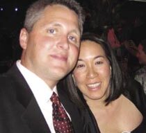 A man in a black suit with a red patterned tie and a woman with shoulder-length hair in a black outfit are smiling and posing for a photo at what appears to be a crowded indoor event. The background is dimly lit, with blurred lights evoking the warm atmosphere of Camp Okizu gatherings.