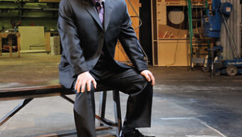 A man in a dark suit and shiny black shoes sits on a wooden bench. The setting appears to be a workshop or backstage area with tools, equipment, and wooden structures in the background. The lighting illuminates Jasson Minadakis, who is looking at the camera and smiling slightly.