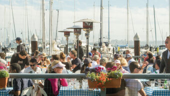A bustling outdoor restaurant filled with people seated at tables covered with blue checkered tablecloths, enjoying their summer eats. Boats and masts are visible in the background, and the sun is shining brightly. The atmosphere appears lively and serene with colorful flowers in the foreground.