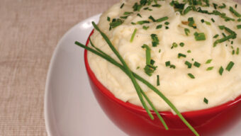 A red bowl filled with creamy mashed potatoes, garnished with chopped chives and whole chive stalks, sits on a white plate against a beige textured background. MMMMM!