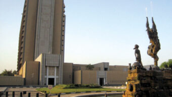 A tall, modern beige building with a unique, angular design stands under a clear sky in the Belvedere area of Baghdad. In the foreground, there is a circular fountain featuring statues, including a figure raising their arms and a smaller figure beside it. The scene appears calm and orderly.