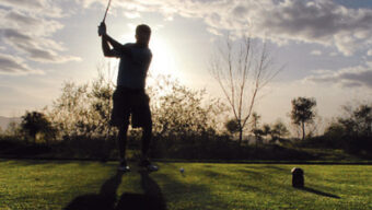 Silhouette of a golfer swinging a club on a golf course with a dramatic sunset or sunrise sky in the background. "He said his drive would be perfect," echoed through trees and foliage surrounding the scene, casting long shadows on the green grass.