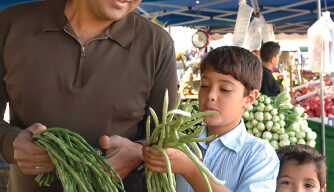 A man and two children are selecting long green beans at an outdoor market. The man, Michael Mina, smiles while holding a bunch of beans. The boy looks curiously at the beans he's holding, and the younger child smiles at the camera. The market stall is filled with various produce.