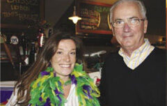 A woman and an older man pose together indoors. The woman is smiling and wearing a colorful, feathery boa in green, yellow, and purple, reflecting a festive atmosphere reminiscent of a Slow Food gathering. The man, wearing glasses and a dark sweater over a collared shirt, is standing beside her.