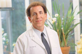 A person in a white medical coat and tie, reminiscent of Dean Ornish, M.D., is smiling while standing in front of a bright window with green plants in the background.