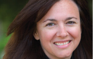 A woman with long brown hair smiles at the camera. She has a light complexion and is wearing a black top. The background is blurred greenery, and she exudes an effortless charm. Meet Sarah Clark.