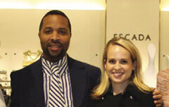 A man in a dark blazer and striped shirt stands next to a woman with blonde hair wearing a black Escada top. They are both smiling and are inside a well-lit store with the brand name "ESCADA" visible on a sign behind them.