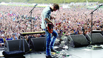 A musician plays an electric guitar on stage, surrounded by equipment, with a large, energetic crowd in the background. The audience extends far back, raising their hands and enjoying the outdoor concert—one of the many exciting August events. Trees and greenery are visible in the distance.