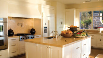 A modern kitchen with white cabinetry, a beige countertop island, recessed lighting, and stainless steel appliances. The island has a faucet and sink, topped with a basket of fruits and vegetables. There's a window on the right, allowing natural light to flood in. This space truly dazzles in the Annual Kitchen Tour.
